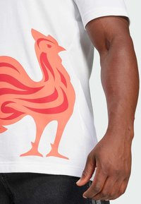 Hvid t-shirt med et stort, stiliseret orange hane grafik med flydende linjer, korte ærmer og en rund halsudskæring.