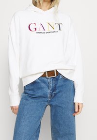 Sudadera blanca con capucha que presenta un colorido texto bordado "GANT AMERICAN SPORTSWEAR", combinada con unos pantalones vaqueros de pierna ancha azul claro y un cinturón marrón.