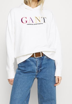 Felpa bianca con cappuccio caratterizzata da un ricamo colorato con la scritta "GANT AMERICAN SPORTSWEAR", abbinata a jeans azzurri a gamba larga e una cintura marrone.