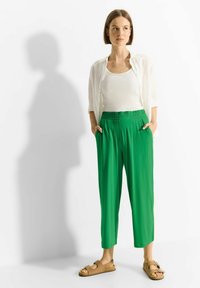 Zielone, luźne spodnie z elastycznym pasem i bocznymi kieszeniami, zestawione z białą koszulą i jasnobrązowymi sandałami. Gładka tekstura materiału.