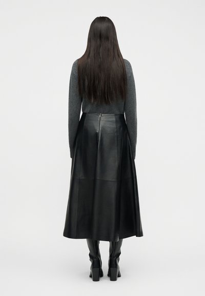 Γυναίκα με μακριά σκουρόχρωμα μαλλιά, που φοράει γκρι πουλόβερ, μαύρη midi δερμάτινη φούστα και μαύρες μπότες με τακούνι, κοιτάζοντας προς τα πίσω σε απλό φόντο.