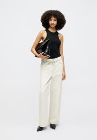 Žena s kučeravými vlasmi, oblečená v bezrukávovom čiernom topu, širokých krémových nohaviciach a čiernych špicatých topánkach, drží veľkú čiernu kabelku pod paží.