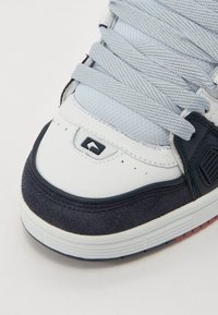 White sneaker featuring navy suede accents, textured leather, and flat grey laces. Toes have a smooth finish with a logo detail and stitched edges.