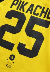 Keltainen puuvillapaita, jossa on mustat numerot "25" ja nimi "PIKACHU". Hihassa on myös musta Poké Ball ja Pikachu -grafiikka helman lähellä.