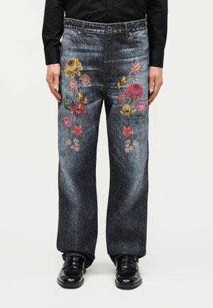 Ανδρικό σκούρο τζιν παντελόνι με πολύχρωμη κεντητή λουλούδια στα μπροστινά μηρά, συνδυασμένο με μαύρα παπούτσια και μαύρο μακρυμάνικο πουκάμισο.