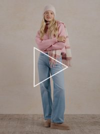 Ružičasti pleteni džemper, svijetloplave široke traperice, bež cipele na platformu, ružičasto-bijeli prugasti šal i odgovarajuća kapa. Minimalistička pozadina.