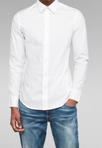 Valkoinen pitkähihaisella napilla varustettu paita, jossa on klassinen kaulus, sileä kangas ja suora helma, yhdistettynä vaaleansinisiin kulutettuihin farkkuihin.