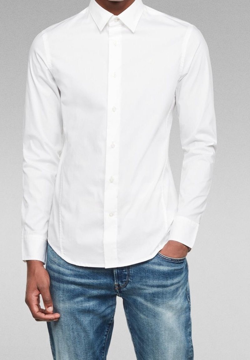 Valkoinen pitkähihaisella napilla varustettu paita, jossa on klassinen kaulus, sileä kangas ja suora helma, yhdistettynä vaaleansinisiin kulutettuihin farkkuihin.