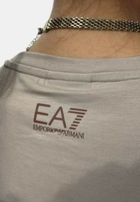 Camiseta de color gris claro con un logotipo bordado en marrón de EA7 y el texto "Emporio Armani" en el cuello. Tejido suave con una textura lisa.