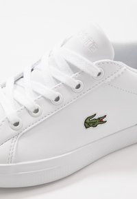 Valkoiset matalat tennarit, joissa valkoiset nauhat, vihreä krokotiililogomainen sivussa ja teksturoitu valkoinen kumipohja.