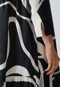 Schwarzes und weißes gemustertes Kleid mit abstrakten Formen, lockerer Passform, kurzen Ärmeln mit Schlitzen und einem gestuften Saum. Aus Baumwollmaterial.