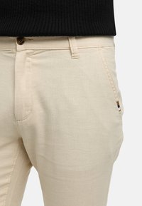 Beige cotton trousers with a smooth texture, featuring a front button closure and subtle side pocket. The design includes fine stitching.