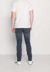 Vitt mönstrat t-shirt med korta ärmar, blå denimjeans och vita sneakers med färgade accenter, sedd bakifrån.