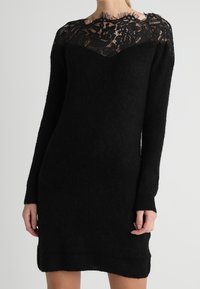 Robe tricotée noire à manches longues, avec un décolleté en dentelle. Texture douce, design ajusté et ourlet droit.