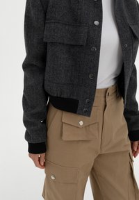 Harmaa herringbone-tyylinen lyhyt takki mustilla hihansuilla ja napeilla, yhdistettynä vaaleanruskeisiin cargo-housuihin, joissa on kaksi etutaskua ja napilla suljettavat läpät.