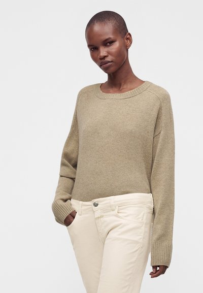 Femme aux cheveux courts, portant un pull en maille beige et un pantalon écru, les mains dans les poches, debout devant un fond clair uni.
