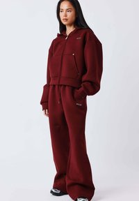 Bordeauxrode zip-up hoodie met capuchon en voorzakken, gecombineerd met bijpassende joggingbroek. Losse pasvorm, zachte textuur en een taille met koord.