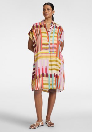 Vrouw draagt een losse, meerkleurige gestreepte jurk tot op de knieën met korte mouwen en witte platte sandalen, staand tegen een effen achtergrond.