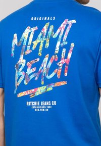 T-shirt en coton bleu avec un graphisme multicolore et le texte "MIAMI BEACH", ainsi qu'un logo de Ritchie Jeans Co. en bas.