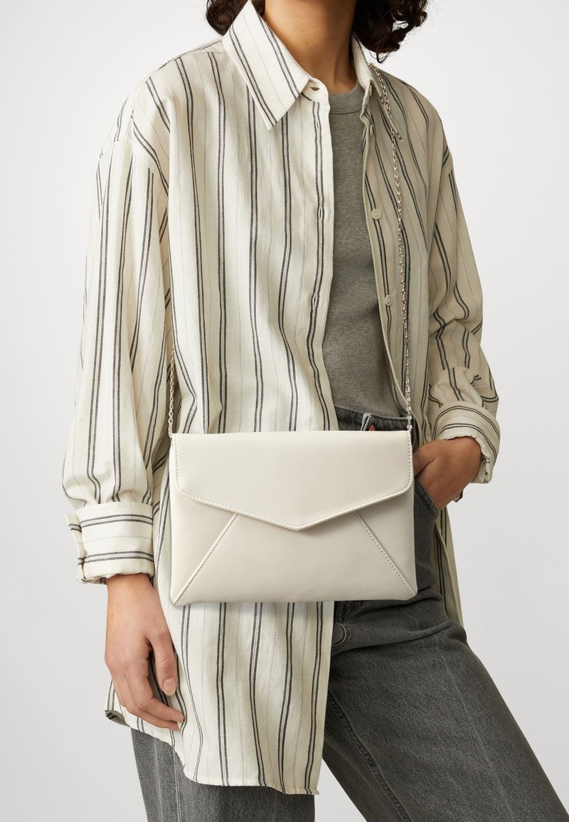 Personne portant une chemise blanche à rayures et un haut gris avec un jean gris, portant un sac bandoulière blanc de style enveloppe.