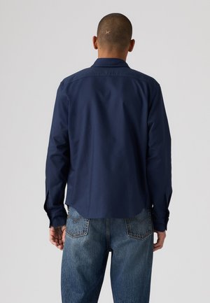 Homme portant une chemise bleu marine à manches longues et un jean bleu, vu de dos sur un fond clair uni.