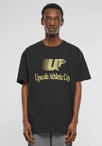 Melna kokvilnas T-kreklā ar dzeltenu grafiku "Upscale Athletic Club", kurā ir skrejceļa figūras dizains un izteikti burti. Parastā piegrieztnē.