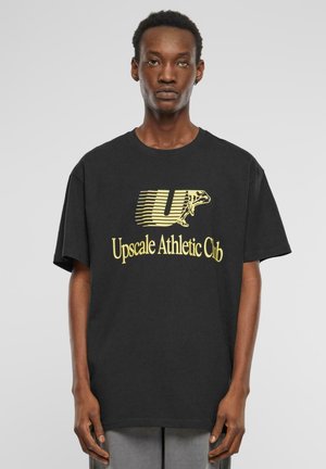 Sort bomulds t-shirt med en gul grafik af "Upscale Athletic Club" med et design af en løbende figur og fed skrift. Standard pasform.