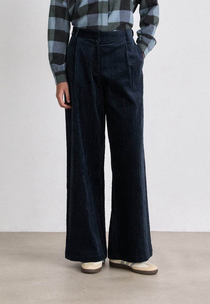 Navy corduroy wide-leg pants with a textured pattern, featuring a high waist and front pleats, paired with cream sneakers.