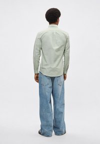 Πράσινο πουκάμισο με μακριά μανίκια από υφασμάτινη υφή, με γιακά που κλείνει με κουμπιά και πιέτα στην πλάτη. Συνδυάζεται με ανοιχτό μπλε τζιν παντελόνι με φαρδιά μπατζάκια.