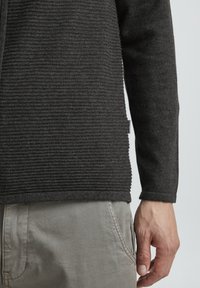 Maglione a coste grigio scuro con maniche lunghe, superficie testurizzata e un piccolo etichetta all'orlo. Indossato con pantaloni grigio chiaro.