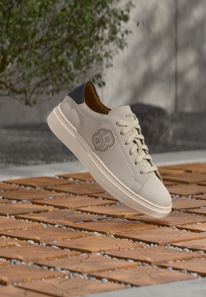 Baskets en daim beige avec languette de talon noire, lacets beiges et logo perforé sur le côté, présentées en suspension au-dessus de carreaux en bois en extérieur.