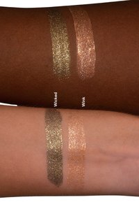 Två metalliska flytande ögonskuggor på hudtonade armar: mörk guld (Wicked) och ljus guld (Wink), båda med glittrande glitterfinish.