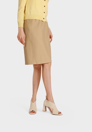 Donna che indossa una gonna beige al ginocchio, un cardigan giallo con bottoni e scarpe beige con tacco a blocco e punta aperta, in piedi su uno sfondo semplice.