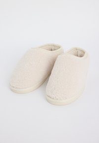 Un par de zapatillas de casa de color crema, peludas, con forro suave y suelas de goma, sobre un fondo blanco.