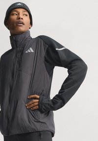 Young person wearing a black Adidas beanie and black and gray Adidas zip-up jacket, standing with one hand on hip, looking to the side.