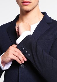 Dark navy blazer with a textured fabric, three black buttons on the cuff, and a single chest pocket. Layered over a white shirt.