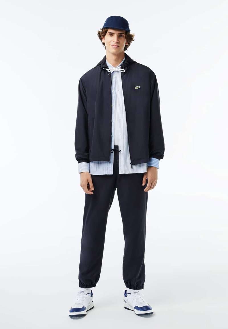 Veste coupe-vent navy avec capuche à cordon, chemise rayée blanche et pantalon sombre. Les accessoires comprennent une casquette navy et des baskets blanches avec des accents bleus.