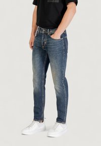 Vaqueros de denim en azul medio con un acabado desgastado, con un diseño de pierna recta, cinco bolsillos y cremallera, combinados con zapatillas blancas.