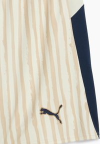 Pantalones cortos deportivos beige con estampado y rayas verticales, y un panel azul marino. Cuenta con un logo de Puma dorado en la parte inferior izquierda. Material texturizado.
