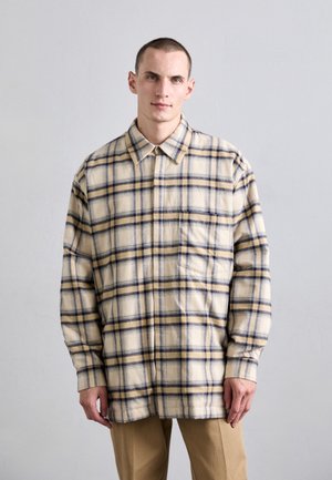 Cămașă flannel cu carouri în nuanțe de bej, navy și galben. Dispune de guler, mâneci lungi și un buzunar pe piept. Textură moale cu o croială lejeră.
