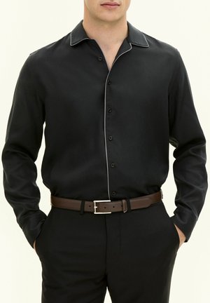 Zwarte blouse met lange mouwen en een spitse kraag, afgewerkt met contrasterende witte piping. Heeft knopen aan de voorkant en een gladde stofstructuur.