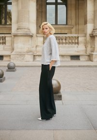 Lyshåret kvinde i grå sweater og sorte bukser med vide ben står med hænderne i lommerne på en byens gade nær stensøjler på en bygning.