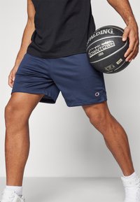 Navyfarbene Mesh-Sportshorts mit elastischem Bund und Logodetails, kombiniert mit einem schwarzen Basketball. Weiße Socken und Turnschuhe.