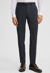 Mann in dunkelblauen, strukturierten Anzughosen, weißem Hemd und schwarzen Lederschuhen, stehend vor einem einfarbigen Hintergrund.