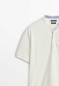 Vit kortärmad polo med texturerad yta, bandkrage, tre knappar fram och ljusblå innerfoder.