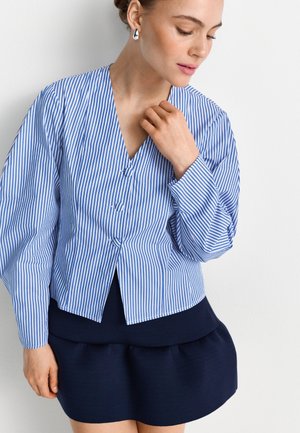 Frau trägt eine blau-weiß gestreifte Bluse mit Knöpfen und langen Ärmeln sowie einen dunkelblauen Rock, blickt nach unten und hält die Hand nahe am Hals.