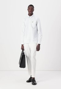 Chemise blanche à boutons avec une poche poitrine et un petit logo, assortie de pantalons blancs et de chaussures en cuir noires, tenant un sac noir.