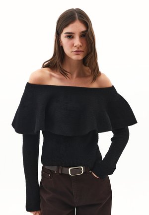 Suéter negro fuera del hombro con un diseño texturizado, mangas largas y un cuello con volantes, combinado con pantalones marrón oscuro y un cinturón.