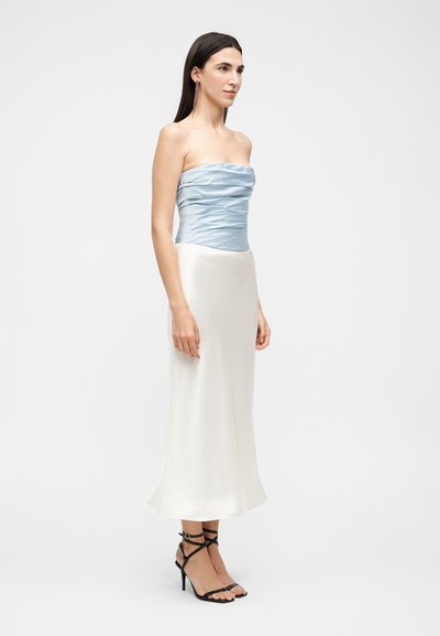 Γυναίκα που φορά ένα αμάνικο, σούρες ανοιχτό μπλε τοπ με μακριά λευκή φούστα και μαύρα σανδάλια με ψηλό τακούνι, στέκεται μπροστά σε απλό φόντο.