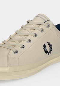Baskets en toile écru avec des lacets beiges, des œillets métalliques et un logo couronne de laurier brodé bleu marine sur le côté.